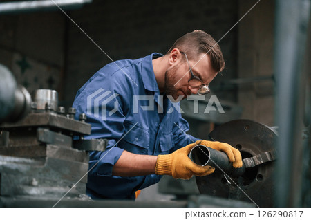 Busy by working with automatic machine. Man at factory and in blue uniform is indoors Busy by working with automatic machine. Man at factory and in blue uniform is indoors 126290817