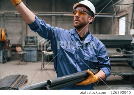 Looking to the side and holding metal pipes. Factory worker in blue uniform is indoors 126290940