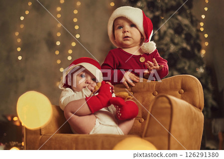Against Christmas tree. Two little kids in Santa hats are celebrating holidays indoors together 126291380