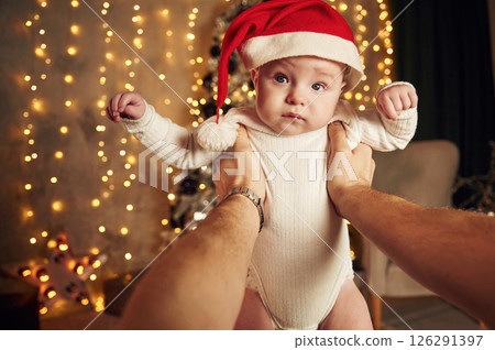 Man is holding a little baby that is in Santa hat. Indoors, with New year festive garlands behind 126291397