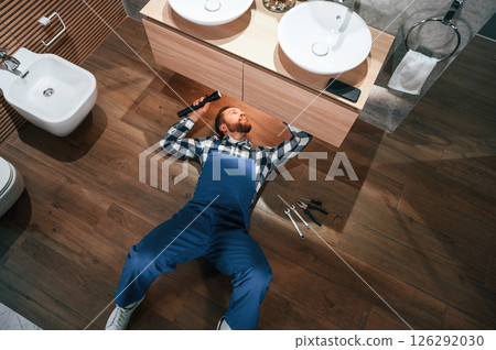 Top view. Lying down on the ground, fixing water tap. Plumber in blue uniform is at work in the bathroom Top view. Lying down on the ground, fixing water tap. Plumber in blue uniform is at work in the bathroom 126292030