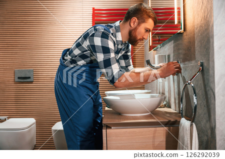 Uses a screwdriver for repairs. Plumber in blue uniform is at work in the bathroom 126292039