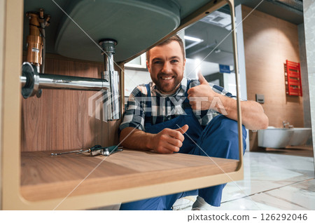 Shwoing thumb up and smiling. Plumber in blue uniform is at work in the bathroom Shwoing thumb up and smiling. Plumber in blue uniform is at work in the bathroom 126292046