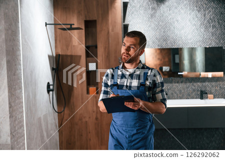 Writes down information in a notebook. Plumber in blue uniform is at work in the bathroom Writes down information in a notebook. Plumber in blue uniform is at work in the bathroom 126292062