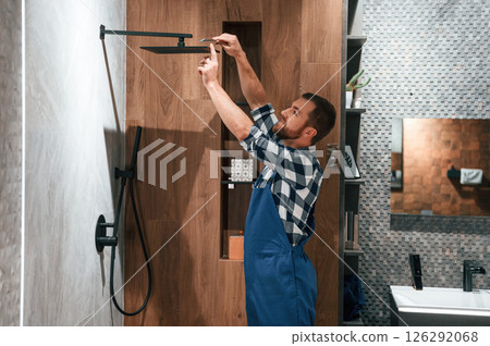 Repairs the shower faucet. Plumber in blue uniform is at work in the bathroom Repairs the shower faucet. Plumber in blue uniform is at work in the bathroom 126292068