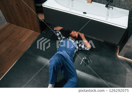 Lying down on the floor, fixing the siphon. Plumber in blue uniform is at work in the bathroom 126292075