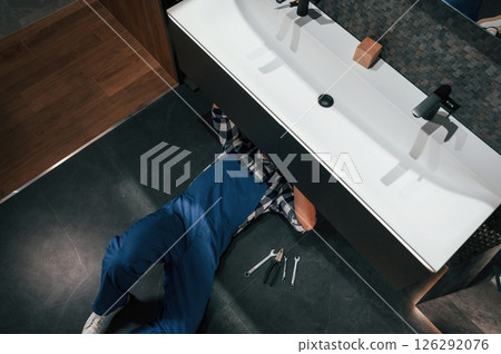 Lying down on the floor, fixing the siphon. Plumber in blue uniform is at work in the bathroom 126292076