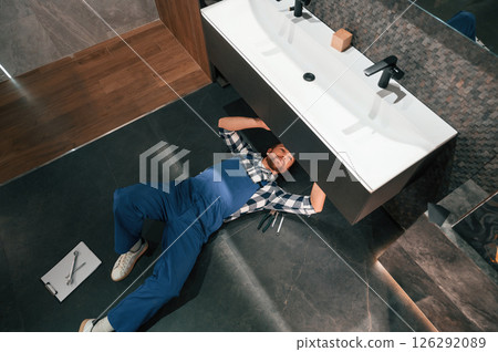 Fixing the siphone. Lying down on the floor. Plumber in blue uniform is at work in the bathroom 126292089