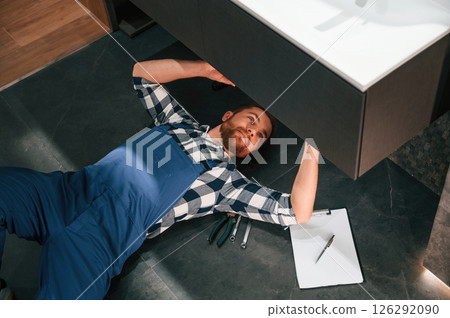 Fixing the siphone. Lying down on the floor. Plumber in blue uniform is at work in the bathroom 126292090