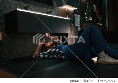 Using flashlight. Fixing water tap. Plumber in blue uniform is at work in the bathroom 126292095