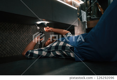 Using flashlight. Fixing water tap. Plumber in blue uniform is at work in the bathroom 126292096