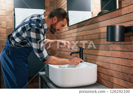 Standing by sink and fixing water tap. Plumber in blue uniform is at work in the bathroom Standing by sink and fixing water tap. Plumber in blue uniform is at work in the bathroom 126292100