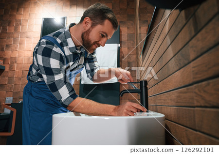 Standing by sink and fixing water tap. Plumber in blue uniform is at work in the bathroom Standing by sink and fixing water tap. Plumber in blue uniform is at work in the bathroom 126292101