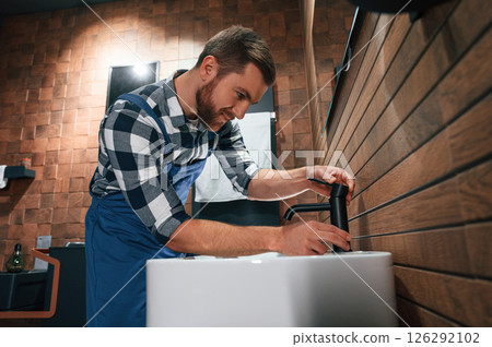 Standing by sink and fixing water tap. Plumber in blue uniform is at work in the bathroom 126292102