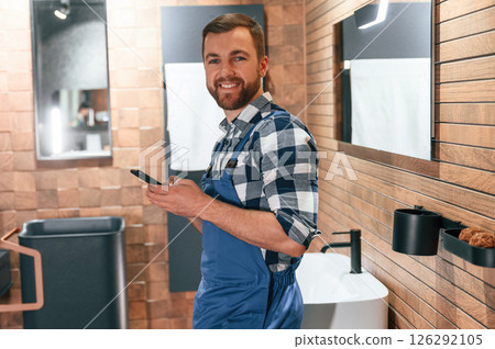 Smiling, holding smartphone in hands. Plumber in blue uniform is at work in the bathroom 126292105