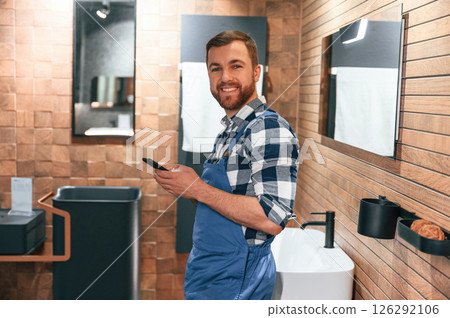 Standing and holding smartphone. Plumber in blue uniform is at work in the bathroom 126292106