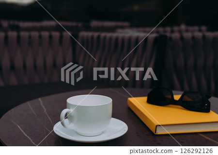 A white cup and a yellow copybook with black sunglasses on a table. A pristine white cup sits next to a yellow book, perfect for relaxation, reading. 126292126