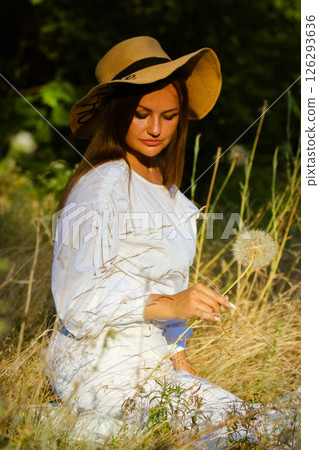 A beautiful gorgeous young woman in a white dress and sun hat poses among tall grass, holding a dandelion, bathed in sunlight. Summertime vacation. 126293636