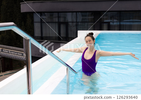 A joyful woman in a purple swimsuit is enjoying a swim in a beautiful outdoor pool. A girl enjoys the outdoor pool, arms wide The modern hotel rooftop 126293640