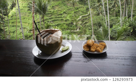Fresh coconut drink and crispy fried snacks on a wooden table overlooking lush green rice terraces with tropical palm trees in a serene vacation setting in Bali 126293973