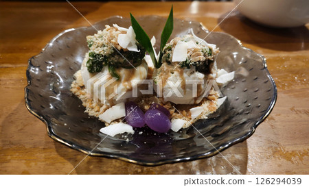 Two scoops of Indonesian-inspired ice cream topped with crushed peanuts, pandan leaves, and coconut flakes, garnished with tapioca pearls, served in a decorative glass bowl. Two scoops of Indonesian-inspired ice cream topped with crushed peanuts, pandan leaves, and coconut flakes, garnished with tapioca pearls, served in a decorative glass bowl. 126294039