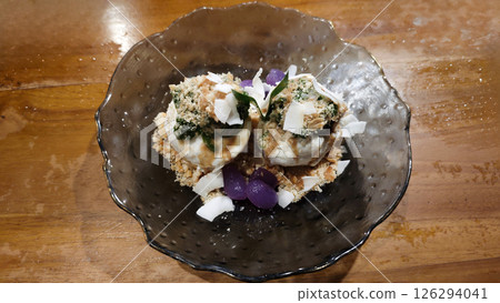 Two scoops of Indonesian-inspired ice cream topped with crushed peanuts, pandan leaves, and coconut flakes, garnished with tapioca pearls, served in a decorative glass bowl. 126294041