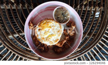 Golden fried egg served with crispy chicken bites and creamy mushroom sauce in a vibrant ceramic bowl, styled on a rustic rattan table, capturing a cozy cafe breakfast ambiance. 126294046