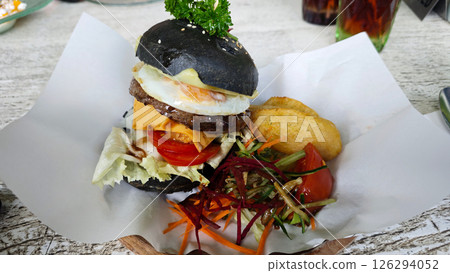 Gourmet black charcoal burger with egg, cheese, and vegetables, served with crispy wedges and fresh salad on parchment, creating a vibrant and appetizing presentation. Gourmet black charcoal burger with egg, cheese, and vegetables, served with crispy wedges and fresh salad on parchment, creating a vibrant and appetizing presentation. 126294052