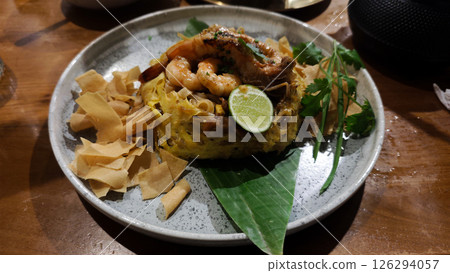 Seafood Char Kway Teow with Egg Net Garnish Served on a Plate with Lime and Crispy Crackers on Wooden Table in a Restaurant Setting 126294057