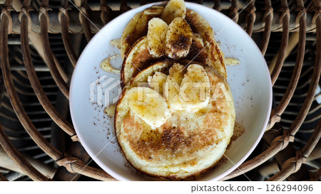 Delicious Banana Pancakes Topped with Sliced Bananas and Drizzled Honey on a White Plate in a Rustic Setting Delicious Banana Pancakes Topped with Sliced Bananas and Drizzled Honey on a White Plate in a Rustic Setting 126294096