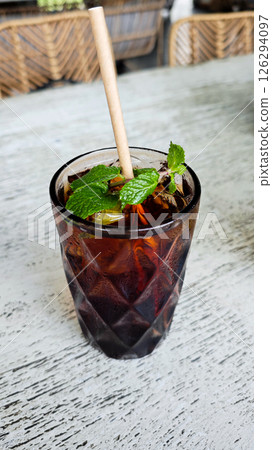Refreshing iced black tea with mint garnish in a textured glass featuring a paper straw on a rustic outdoor cafe table setting for a relaxing beverage break Refreshing iced black tea with mint garnish in a textured glass featuring a paper straw on a rustic outdoor cafe table setting for a relaxing beverage break 126294097
