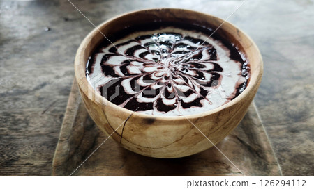 Elegant bowl of black glutinous rice porridge drizzled with intricate coconut milk art, served in a rustic wooden bowl on a textured wooden surface for a traditional Indonesian dessert experience. Elegant bowl of black glutinous rice porridge drizzled with intricate coconut milk art, served in a rustic wooden bowl on a textured wooden surface for a traditional Indonesian dessert experience. 126294112