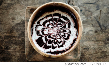 Elegant bowl of black glutinous rice porridge drizzled with intricate coconut milk art, served in a rustic wooden bowl on a textured wooden surface for a traditional Indonesian dessert experience. Elegant bowl of black glutinous rice porridge drizzled with intricate coconut milk art, served in a rustic wooden bowl on a textured wooden surface for a traditional Indonesian dessert experience. 126294113