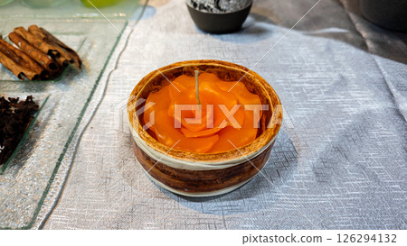 Orange flower-shaped candle in a rustic ceramic bowl displayed on a table with natural elements and spices in a cozy setting 126294132
