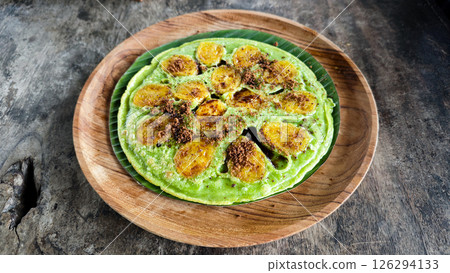 Vibrant green banana pancake with caramelized banana slices and brown sugar, served on a wooden plate with a rustic dark wood background 126294133