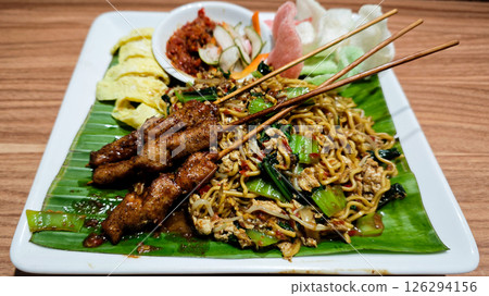 Stir-fried noodles served with grilled chicken skewers, omelette slices, and assorted vegetables on a banana leaf-lined plate with sambal and crackers on the side, presented on a wooden table 126294156