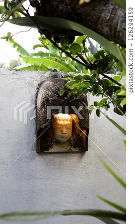 Illuminated stone Buddha head sculpture in garden surrounded by green leaves and tree branches against white wall background 126294159