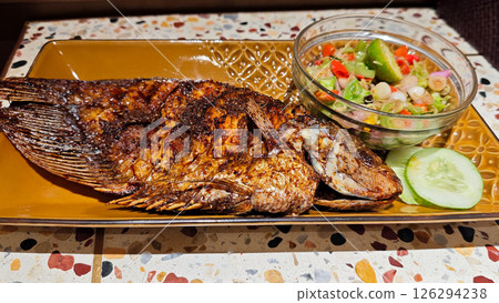 Close-up of Grilled Gourami Fish Served with Balinese Sambal Matah and Fresh Cucumber Garnish on a Traditional Brown Plate in a Restaurant with Terrazzo Table Background Close-up of Grilled Gourami Fish Served with Balinese Sambal Matah and Fresh Cucumber Garnish on a Traditional Brown Plate in a Restaurant with Terrazzo Table Background 126294238
