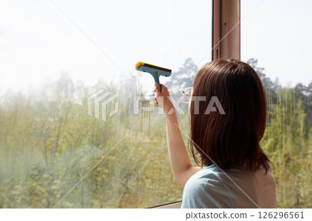 A young women is cleaning a large window with a squeegee, revealing a scenic outdoor view. A young women is cleaning a large window with a squeegee, revealing a scenic outdoor view. 126296561