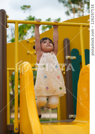 happy toddler girl hanging the yellow bar by her hand at outdoor playground in park 126297538