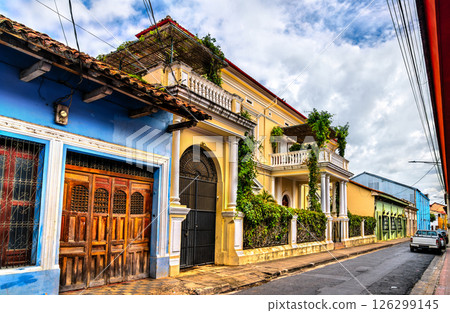 Row of vibrant colonial-style houses with ornate balconies, arched windows, and lush greenery lines a narrow cobblestone street in Granada, Nicaragua. The pastel colors and architectural details 126299145