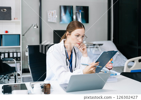 Confident young Caucasian female doctor in white medical uniform sit at desk working on computer. Smiling use laptop write in medical journal in clinic. 126302427