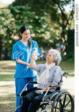 Asian physiotherapist helping elderly woman patient stretching arm during exercise correct with dumbbell in hand during training hand with patient Back problems in the garden. 126302677