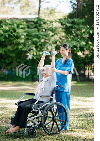 Asian physiotherapist helping elderly woman patient stretching arm during exercise correct with dumbbell in hand during training hand with patient Back problems in the garden. 126302712