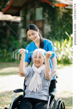 Asian physiotherapist helping elderly woman patient stretching arm during exercise correct with dumbbell in hand during training hand with patient Back problems in the garden. 126302714