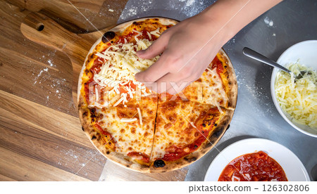 A hand sprinkles shredded cheese on a sliced cooked pizza, with bowls of ingredients nearby. 126302806