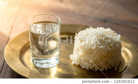 A glass of water and a mound of white rice on a brass plate, illuminated by warm light. A glass of water and a mound of white rice on a brass plate, illuminated by warm light. 126302812
