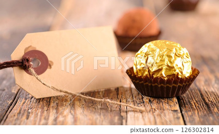 A gold wrapped chocolate praline with a gift tag on a rustic wooden table. A gold wrapped chocolate praline with a gift tag on a rustic wooden table. 126302814