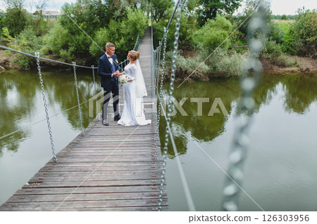 Beautiful newlywed couple in love posing on suspension bridge against summer nature background Beautiful newlywed couple in love posing on suspension bridge against summer nature background 126303956