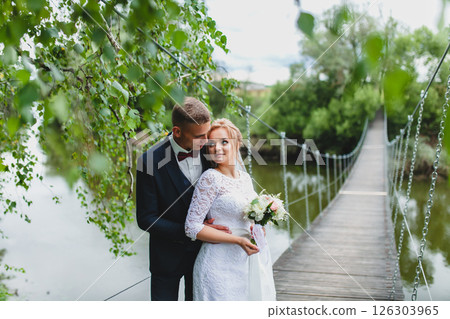 Beautiful newlywed couple in love posing on suspension bridge against summer nature background Beautiful newlywed couple in love posing on suspension bridge against summer nature background 126303965
